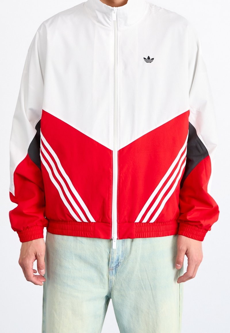 Red and white track jacket with a high collar, zip closure, chevron design, and three white stripes on each sleeve. Black accents on sides.