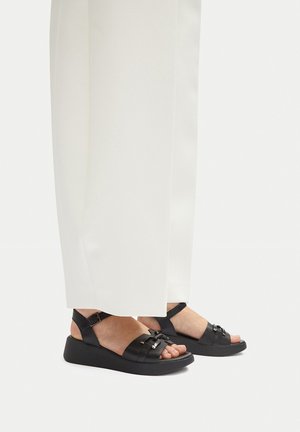 Persona che indossa pantaloni bianchi e zeppe nere aperte con cinturini alla caviglia e nodo decorativo sulle cinghie anteriori.