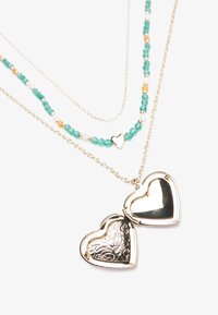 Guldtonet hjerteformet locket vedhæng med indviklet blomsterdesign, parret med lagdelte kæder i turkise og perler. Justerbar længde.