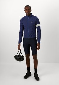 Veste de cyclisme bleue marine avec zip frontal et accent blanc, associée à un short foncé, des chaussures de cyclisme noires et un casque tenu à la main.