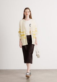 Cardigan crème avec des rayures jaunes sur un t-shirt blanc, associé à une jupe crayon marron et des baskets grises. Sac bandoulière simple.