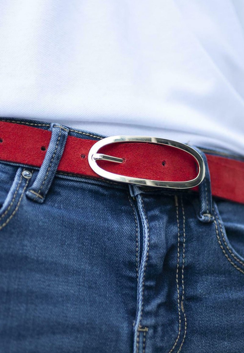 Cinturón de ante rojo con una hebilla ovalada de plata, llevado sobre jeans de mezclilla azul, con costuras visibles y detalles de botones.
