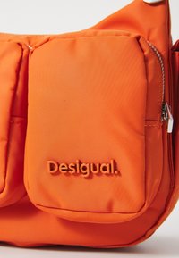 Pomarańczowa torba nylonowa z wyraźną przednią kieszenią, zamknięciem na zamek oraz wytłoczonym logo "Desigual". Gładka tekstura i smukły design.