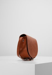 Borsa a tracolla in pelle marrone con chiusura a pattina e tracolla regolabile posizionata su una superficie bianca contro uno sfondo uniforme.