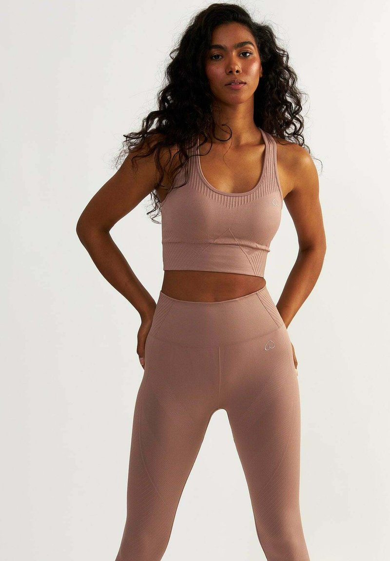 Svetle růžová sportovní souprava obsahující těsný crop top s jemně žebrovanou texturou a legíny s vysokým pasem, které mají decentní logo srdce a akcenty vzoru.