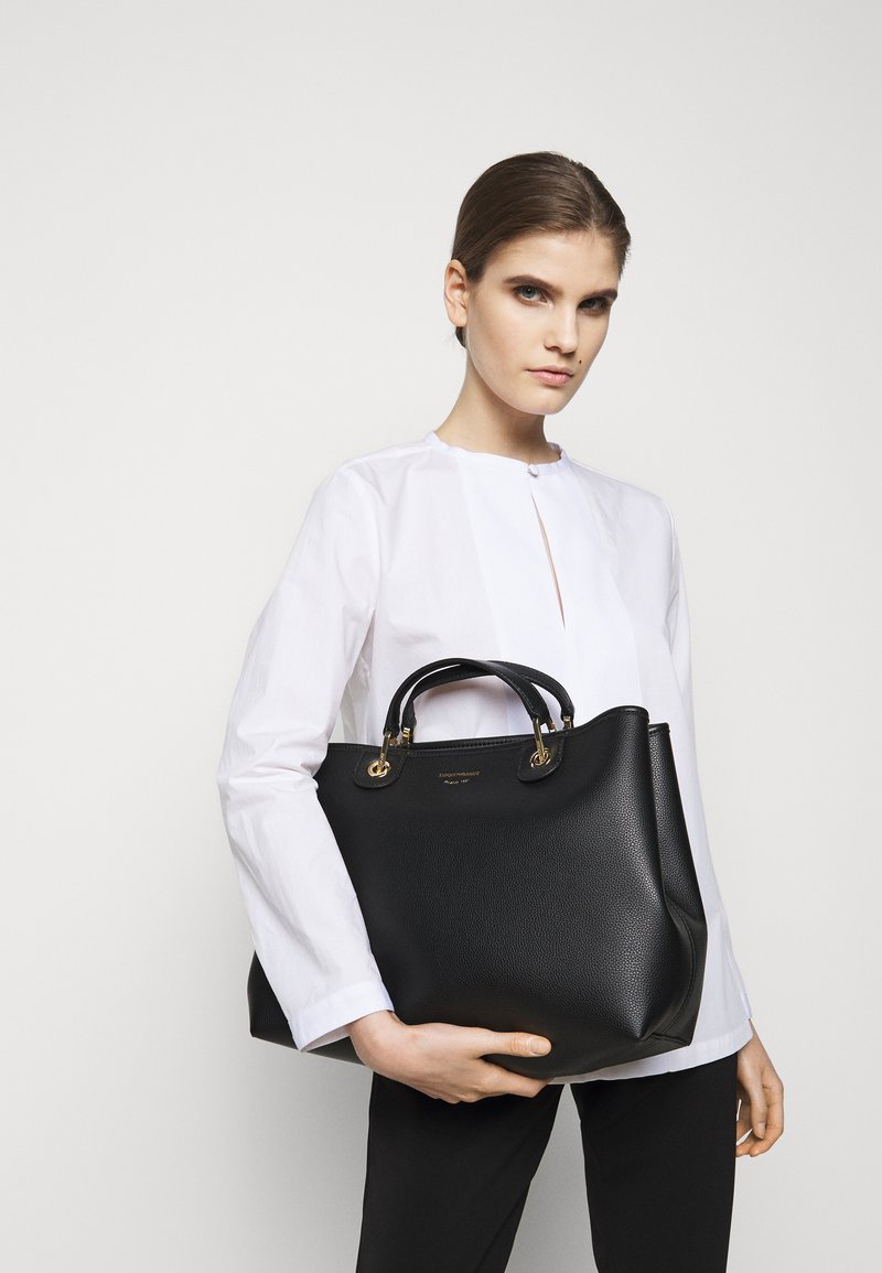Borsa in pelle nera con hardware dorato, forma strutturata, superficie testurizzata, manici superiori e logo centrato sulla parte anteriore.