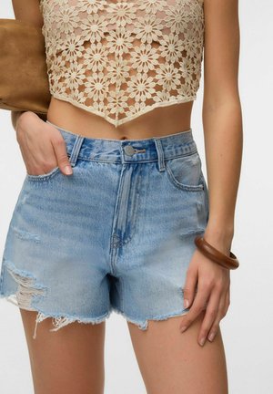 Kvinde iført lyseblå slidte denimshorts og en beige blomsterhæklet croptop med et brunt armbånd om håndleddet.