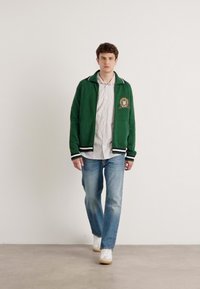 Groene zip-up jas met gestreepte kraag, embleem op de borst, gedragen over een gestreept shirt, in combinatie met lichtblauwe jeans en witte sneakers.