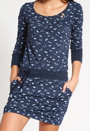 Frau trägt ein marineblaues langärmeliges Kleid mit hellblauen Vogelmustern, zwei Knöpfen nahe dem Ausschnitt und den Händen in den Rocktaschen.