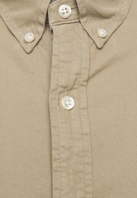 Beżowa koszula zapinana na guziki z wywiniętym kołnierzykiem, z białymi plastikowymi guzikami i teksturowanym materiałem. Szczegóły przeszycia widoczne wzdłuż listwy guzika.