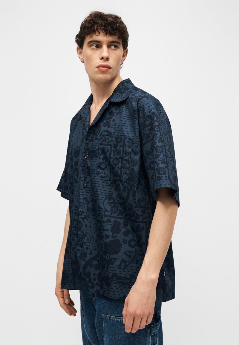 Giovane uomo con capelli ricci che indossa una camicia a maniche corte blu scuro con motivi e jeans blu, che guarda di lato.