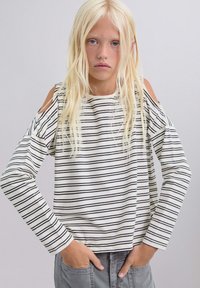 Niña rubia con pelo largo, vestida con una blusa de manga larga de rayas blancas y negras con los hombros al descubierto y pantalones grises, con las manos en los bolsillos.