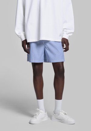 Blue and white striped shorts made of lightweight fabric, featuring two side pockets, worn with a white oversized sweatshirt and white sneakers.