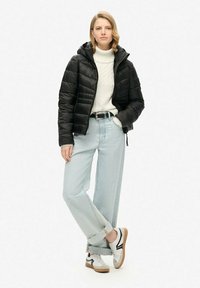 Zwarte gewatteerde pufferjack met capuchon, crèmekleurige kabelgebreide trui, lichtblauwe wijde jeans en witte sneakers met zwarte accenten.