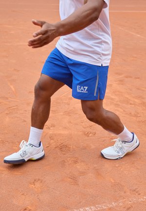 Blue athletic shorts with a logo, paired with white sneakers featuring gray and green accents, on a clay tennis court.