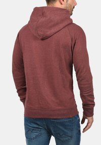 Maroon hooded sweatshirt with a textured finish, ribbed cuffs, and hem, featuring a stitched logo tag on the lower side.