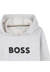 Sweat à capuche gris avec une texture douce, présentant un grand logo "BOSS" noir sur le devant. La couture intérieure révèle un doublage beige avec de fines rayures noires et dorées.