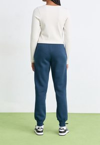 Valkoinen pitkähihaisten ribbineuletakki, joka on yhdistetty löysiin tummansinisiin collegehousuihin. Mustat ja valkoiset urheilukengät logolla. Vihreä lattia taustalla.