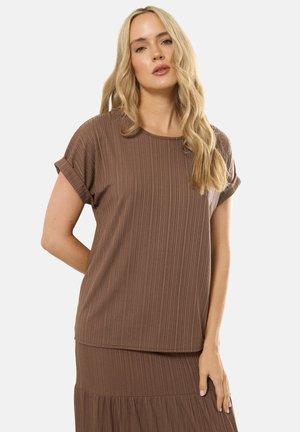 Jonge vrouw met lang blond haar, gekleed in een bruin geribd topje met korte mouwen en een bijpassende rok, staand voor een effen achtergrond.