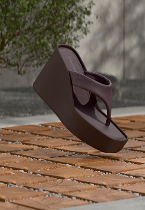 Tamsiai ruda Calvin Klein platforminė basutė, sklendžianti virš medinės ir žvyro rašto lauko grindų, su žaliomis augmenijos fone.