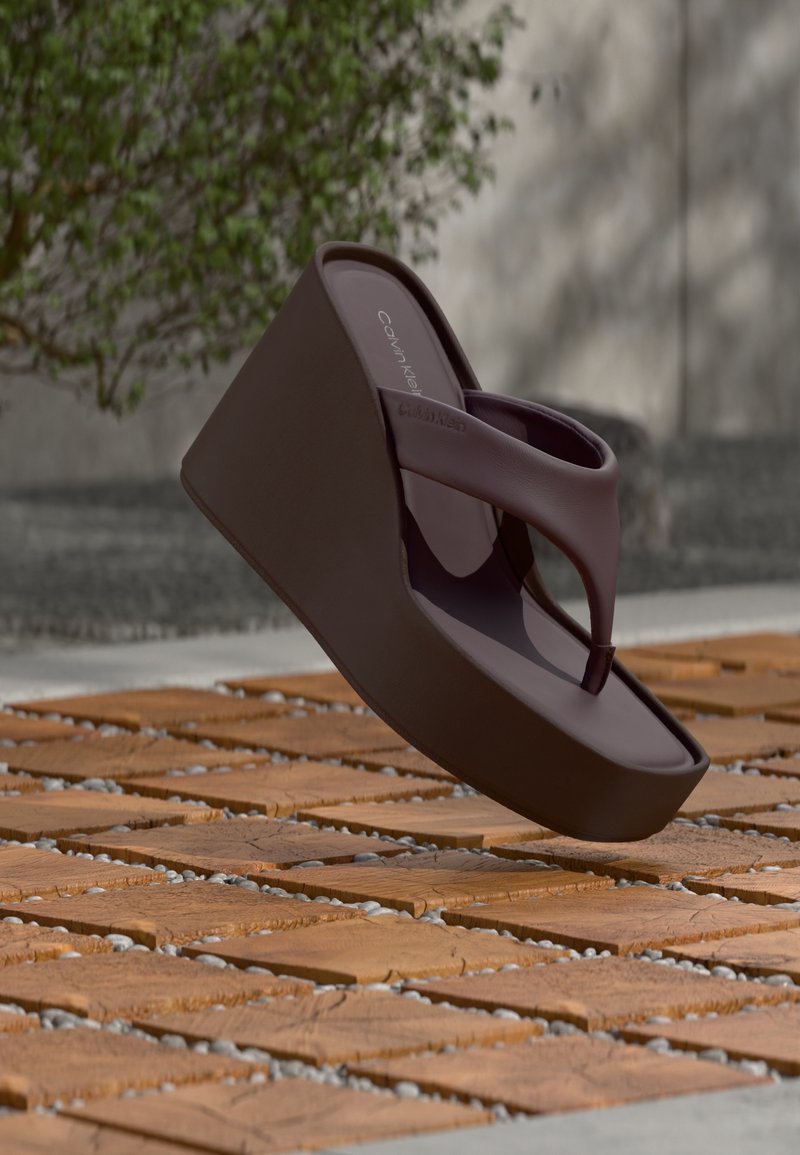 Σκούρο καφέ πλατφόρμα σανδάλι Calvin Klein που αιωρείται πάνω από ξύλινο δάπεδο με σχέδιο από χαλίκι, με φόντο φυτά.