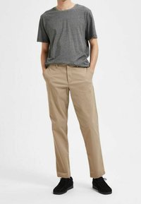 Grå t-shirt med kort ärm, beige raka byxor, svarta skor. Byxorna har framfickor och en gylf framtill med knappstängning.