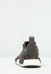 Dark brown athletic shoe with a knit upper, textured accents, and a white foam sole. Features a pull tab with brand logo on the heel.