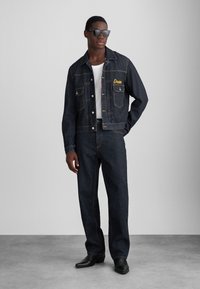 Chaqueta de mezclilla oscura con dos bolsillos delanteros, logo amarillo bordado "Dean", combinada con pantalones de mezclilla oscuros. Modelo con gafas de sol negras.