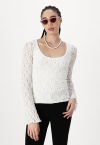 Weißes Spitzenoberteil mit langen, ausgestellten Ärmeln und einem Rundhalsausschnitt. Kombiniert mit schwarzen Hosen, accessoriert mit einer Perlenkette und rosa Sonnenbrille.