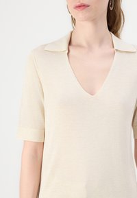Top beige a maniche corte con scollo a V e colletto classico. Il tessuto appare leggero con una texture liscia.