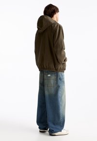 Chaqueta con capucha de color verde oliva con dobladillo elástico y bolsillos laterales, combinada con vaqueros de mezclilla azul claro de pierna ancha con doble bolsillo trasero.