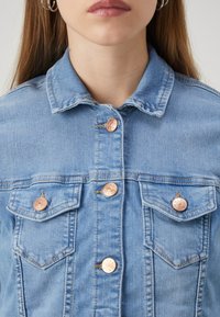 Ljusblå denimjacka med klassisk krage, prydd med kopparfärgade knappar och två bröstfickor med sömnadsdetaljer.