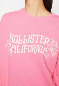 Rosa fleece-tröja med vit grafisk text "HOLLISTER CALIFORNIA" och illustrationer av blommor och en svamp på framsidan.