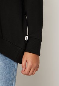 Felpa nera con polsini e orlo a costine, con una piccola etichetta bianca Vans. Jeans blu in denim visibili sotto la felpa.