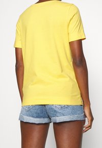 Camiseta de algodón amarilla con mangas cortas y escote redondo, combinada con pantalones cortos de mezclilla. Costuras visibles y ligera textura en la tela.