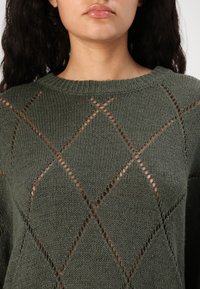 Grüner Strickpullover mit lockerer Passform, rautenförmigen Ausschnitten und einem gerippten Kragen. Die Textur ist weich und hat ein leicht grobmaschiges Strickmuster.