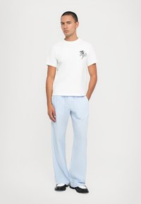 Man standing with hands in pockets wearing white graphic t-shirt, light blue striped wide-leg pants, and black-and-white shoes.