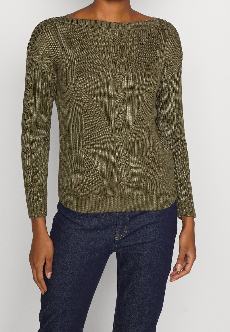 Personne portant un pull torsadé vert olive à manches longues associé à un jean bleu foncé sur un fond uni.