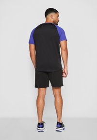 Svart og lilla treningstrøye med korte ermer, med en svart kropp og lilla raglan-ermer. Kombinert med svarte shorts og sko.