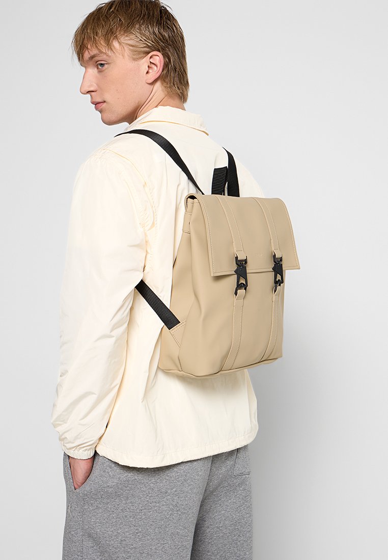 Zaino beige con un design strutturato, spallacci neri regolabili e un coperchio superiore fissato da due fibbie. Materiale liscio con cuciture sottili.