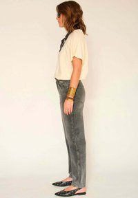 Haut à manches courtes beige clair, jean noir délavé, chaussures slip-on noires avec accents argentés, et bracelets dorés empilés portés au poignet.