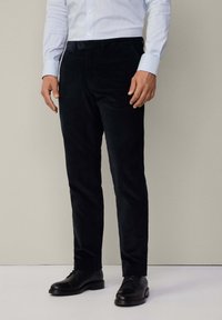 Pantaloni in corduroy scuro, slim fit, con una texture liscia, dotati di chiusura a bottone e tasche frontali, abbinati a scarpe nere.