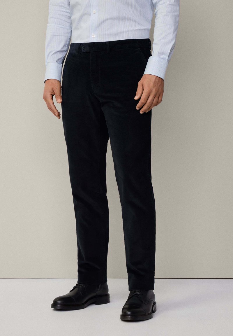 Pantaloni in corduroy scuro, slim fit, con una texture liscia, dotati di chiusura a bottone e tasche frontali, abbinati a scarpe nere.