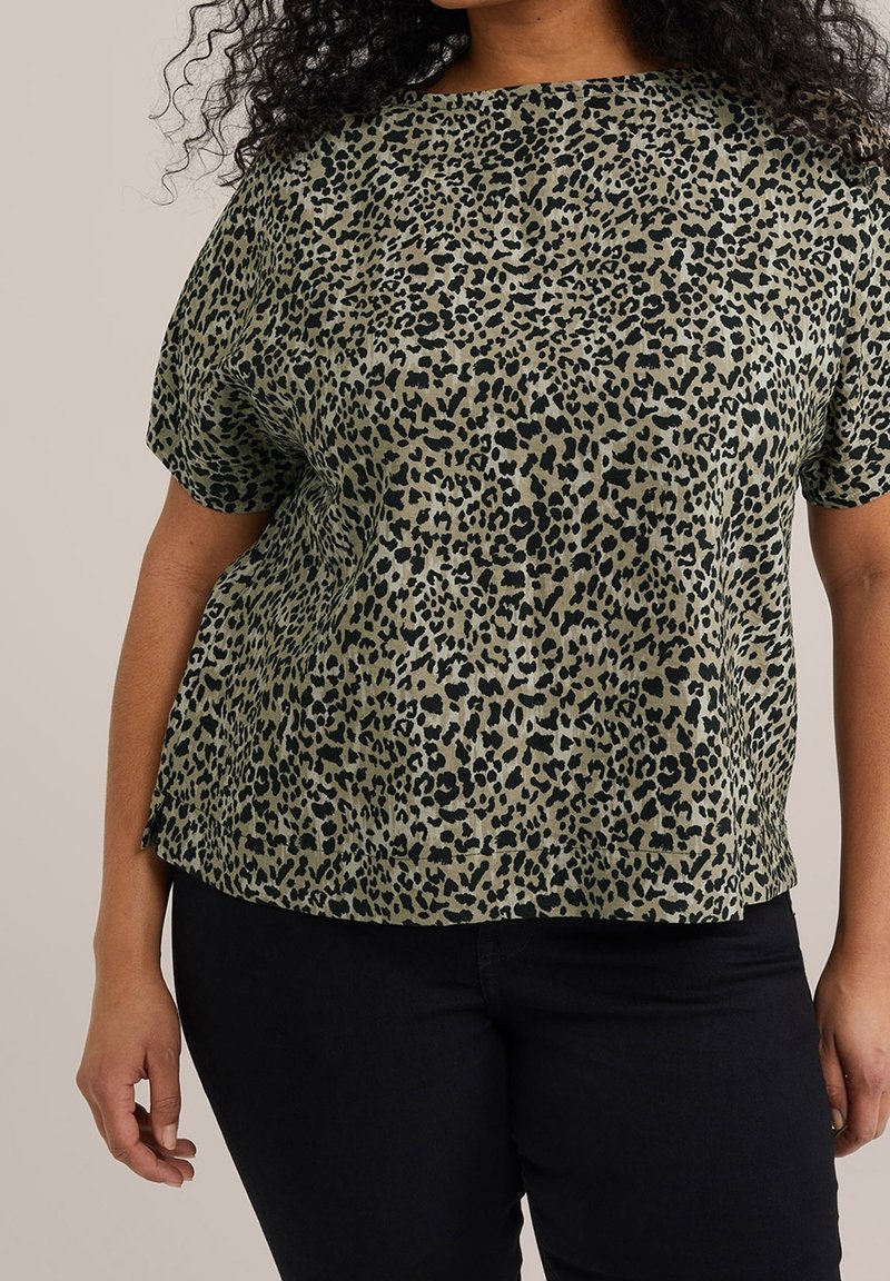 Femme portant un chemisier à manches courtes imprimé léopard vert et noir avec un pantalon noir, vue des épaules aux hanches.