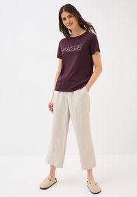 T-shirt en coton violet avec un graphique "ALOHA", associé à un pantalon large en lin beige clair et des chaussures slip-on beiges.