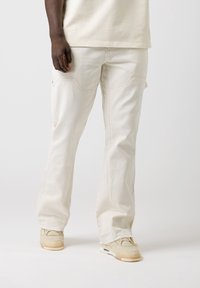 Pantalons cargo beiges à coupe ample, avec poches avant, détails cousus et design à jambe droite. Portés avec des baskets de couleur claire.