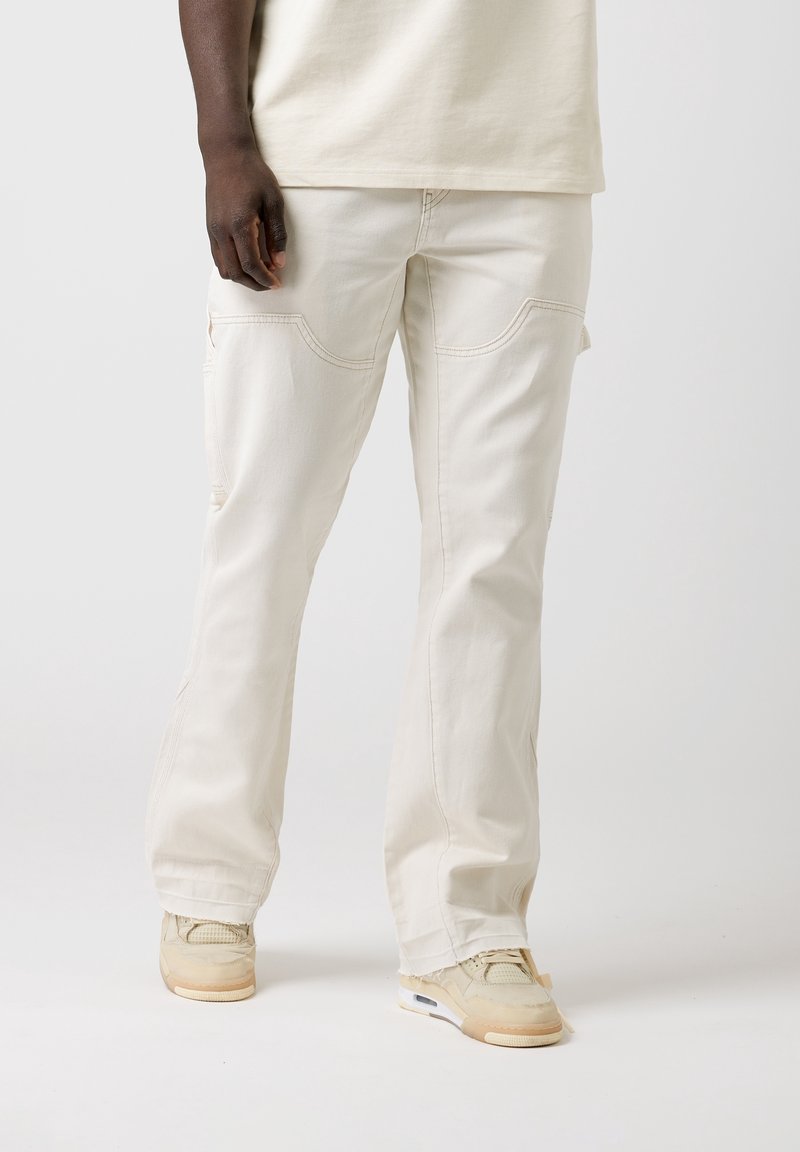 Pantalons cargo beiges à coupe ample, avec poches avant, détails cousus et design à jambe droite. Portés avec des baskets de couleur claire.