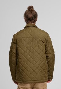 Oliwkowa pikowana kurtka z diamentowym wzorem, z zapięciami na guziki oraz detalem przy kołnierzu. Tekstura materiału wydaje się gładka i uporządkowana.