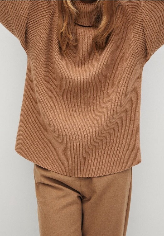 Personne portant un pull à col roulé côtelé marron et un pantalon marron assorti, les bras levés au-dessus de la tête sur un fond uni.
