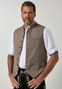 Hombre vistiendo un chaleco marrón abotonado sobre una camisa blanca remangada y pantalones tradicionales negros bordados, de pie con las manos entrelazadas.